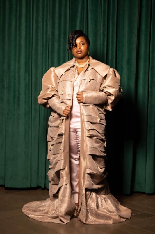 Person in elaborate gown against green curtain.